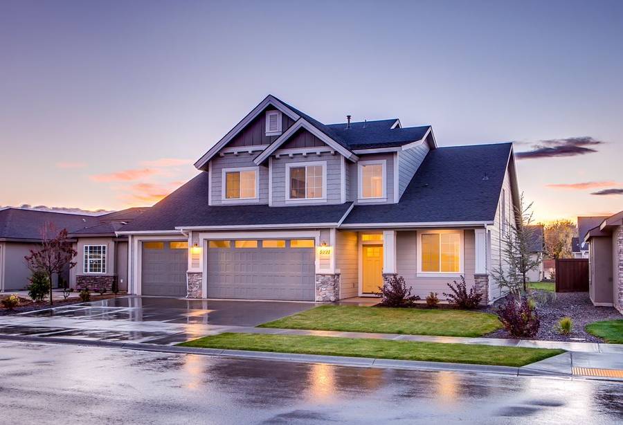 A two-story home at dusk with warm lights on and a wet driveway after an evening rain